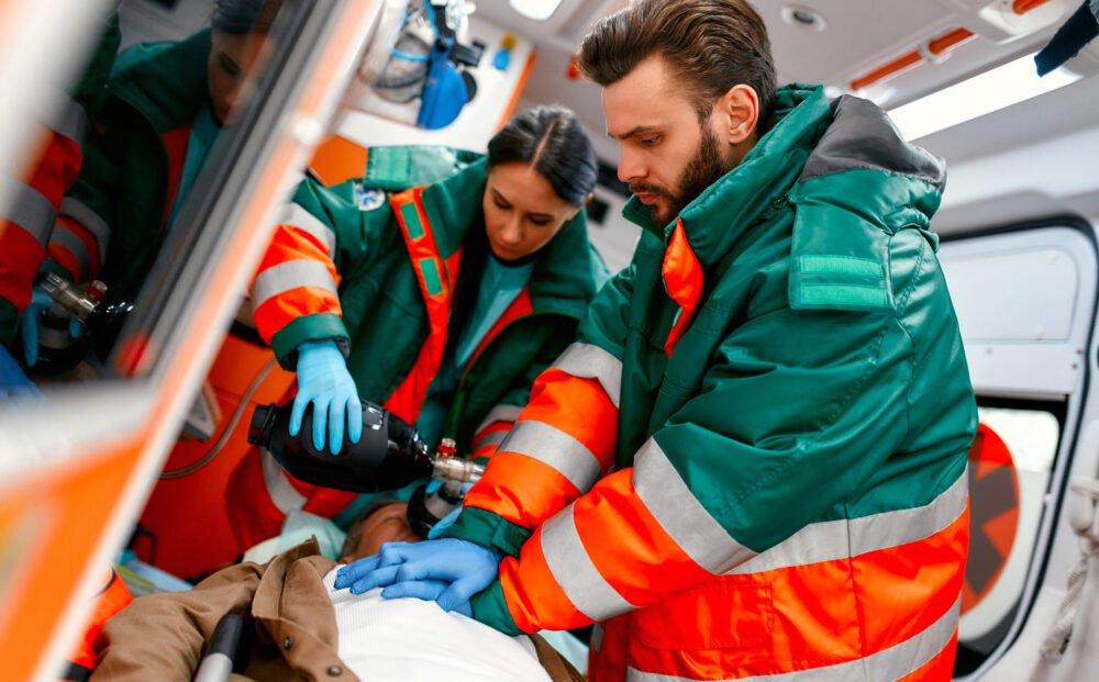 Two paramedics are resuscitating a senior lying on a gurney in an ambulance by performing chest compressions and connecting to a ventilator.