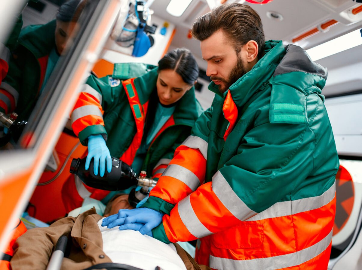 Two paramedics are resuscitating a senior lying on a gurney in an ambulance by performing chest compressions and connecting to a ventilator.
