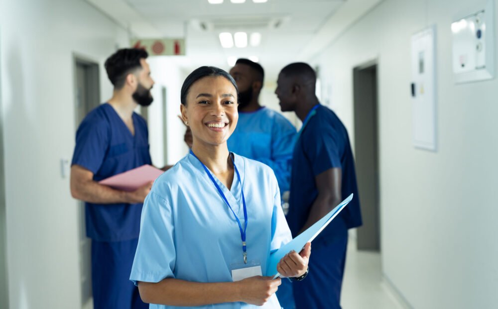 Portrait of smiling biracial female healthcare worker with file in busy hospital corridor. Hospital, medical and healthcare services, copy space.