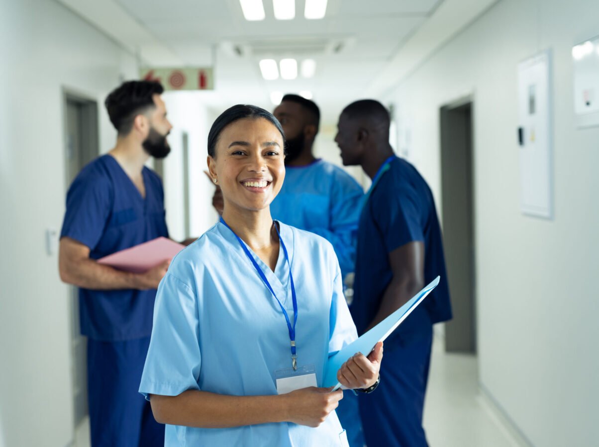 Portrait of smiling biracial female healthcare worker with file in busy hospital corridor. Hospital, medical and healthcare services, copy space.