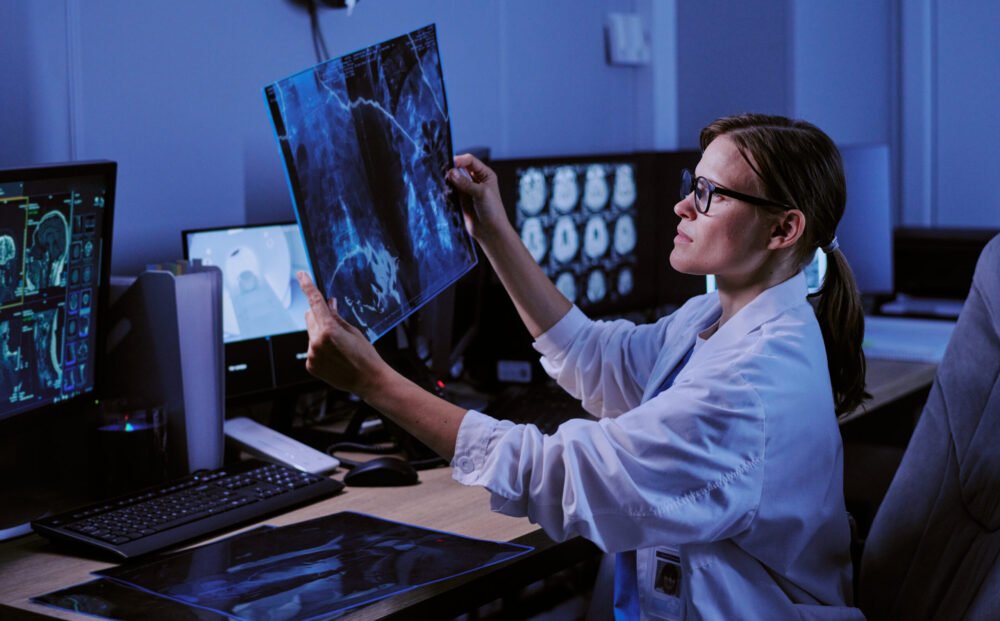 Caucasian young adult woman doctor examining X-ray scan at workstation in medical imaging center, analyzing radiology images on computer monitors, wearing eyeglasses and lab coat